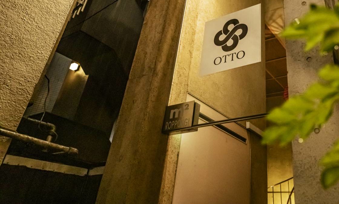 雨の夜、ビルの看板塔に灯る白い四角いパネル。黒い結び目のようなマークと「OTTO」の文字。その下に「m² ROPPONGI」のプレート。