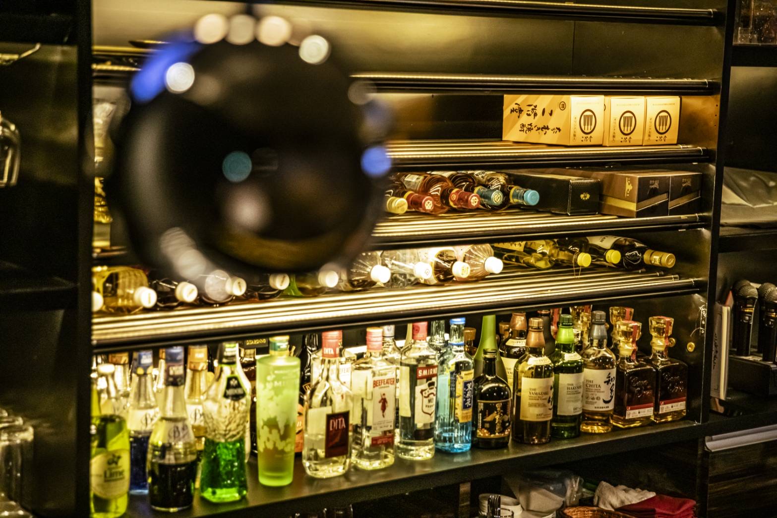 照明に照らされたバックバーの棚。寝かせたボトルと並んだ酒瓶、手前にぼけた球形のペンダントライト。