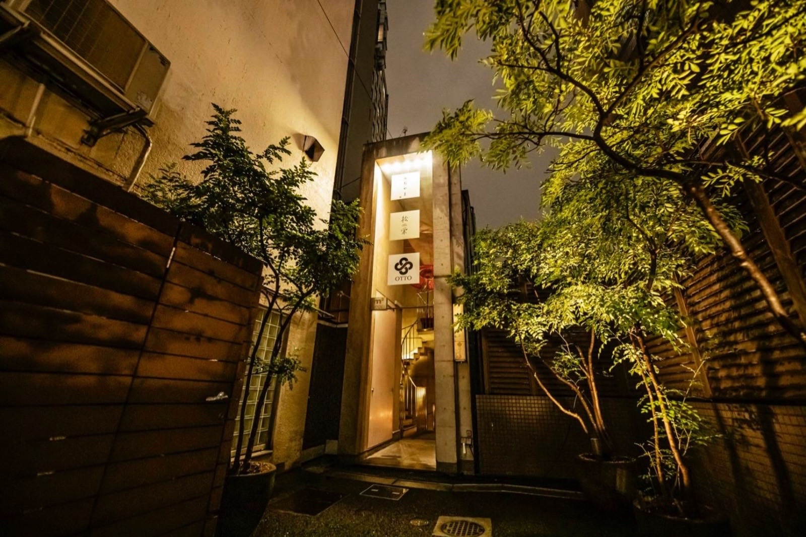 夜、木の塀とライトアップされた木々の間を抜けた先に立つコンクリートの看板塔。いちばん下の白いパネルに黒い結び目のマークと「OTTO」。明るい入口の奥にらせん階段。